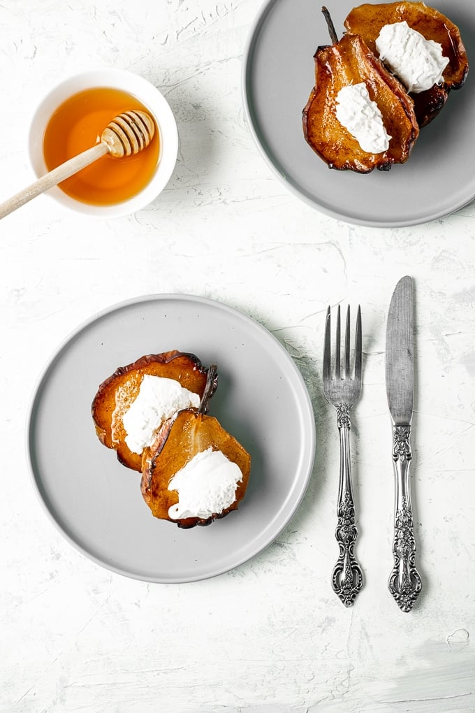 Roasted pears topped with clotted cream served on two grey round plates and honey in a bowl, a fork and a knife on the side.