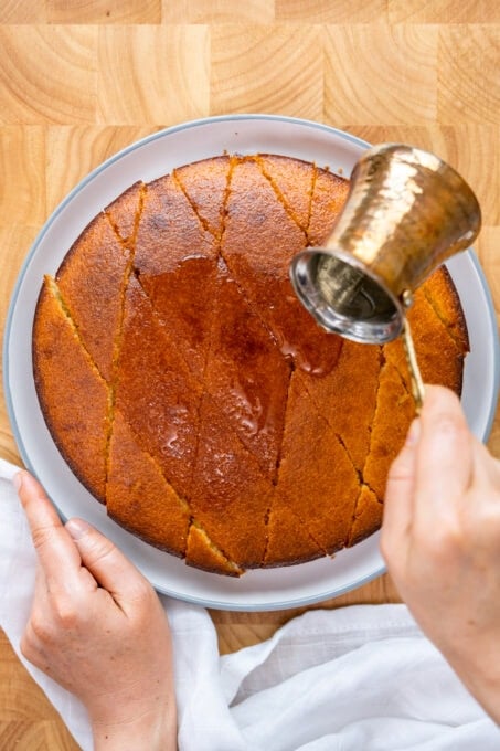 Hands pouring sugar syrup in a copper coffee pot over the cooked semolina cake.