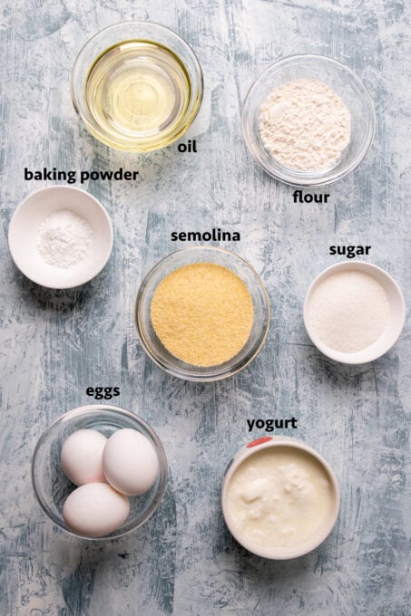 Semolina, flour, eggs, sugar, baking powder, oil and yogurt all in separate bowls on a light background.