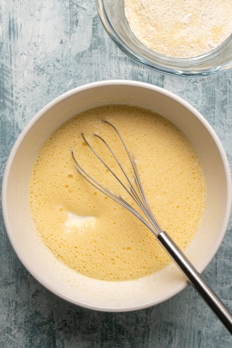 Yogurt and oil added in the egg sugar mixture in a white bowl and a hand whisk inside it.