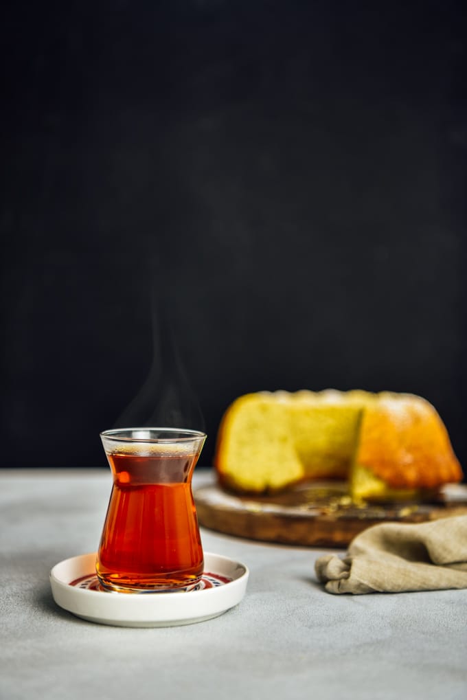Hot Turkish tea served in a traditional Turkish tea glass on a tea plate photographed from front view with a dark background. Accompanied by a lemon cake.