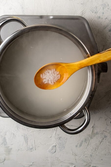Rice cooking in simmering water in a pot and a wooden spoon showing the rice over the pot.