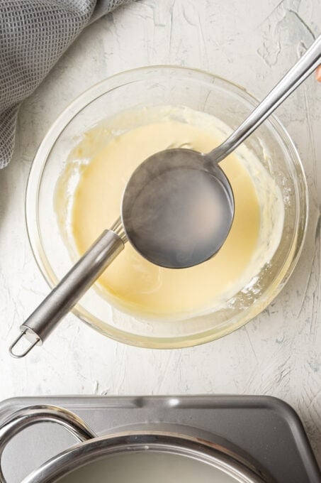 Tempering the yogurt mixture in a glass bowl with a ladle of hot water.