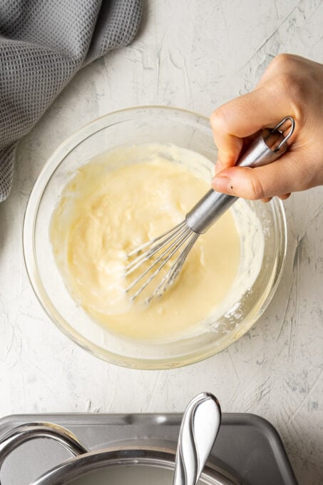A hand whisking yogurt mixture in a glass bowl to temper.