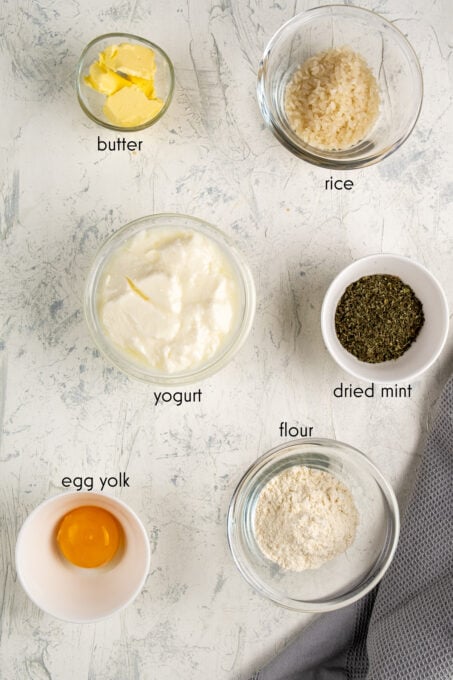 Yogurt, egg yolk, flour, dried mint, rice and butter in separate bowls on a white background.