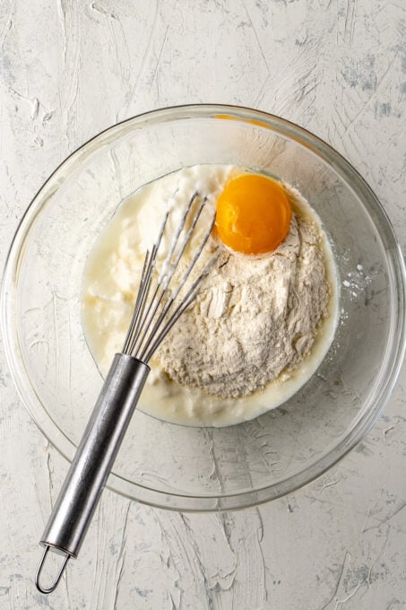 Yogurt, flour and egg yolk in a glass bowl and a hand whisk in it.