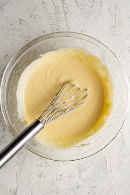 Yogurt, flour and egg yolk mixture in a glass bowl and a hand whisk in it.