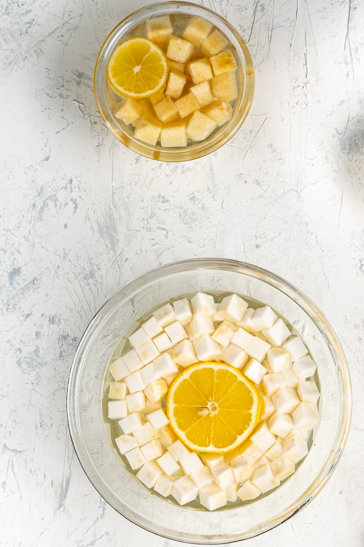 Diced celeriac and diced quince topped with lemon slice in two separate bowls filled with water.