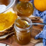 Pumpkin jam in a jar with a spoon in it, some jam slathered on bread slices on the side.