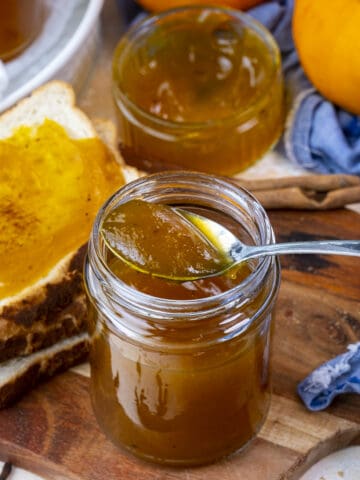 Pumpkin jam in a jar with a spoon in it, some jam slathered on bread slices on the side.