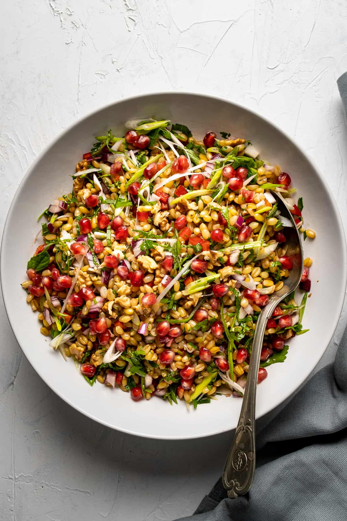 Wheat berries, herbs, onions and pomegranate arils combined in a white bowl and a spoon in it.