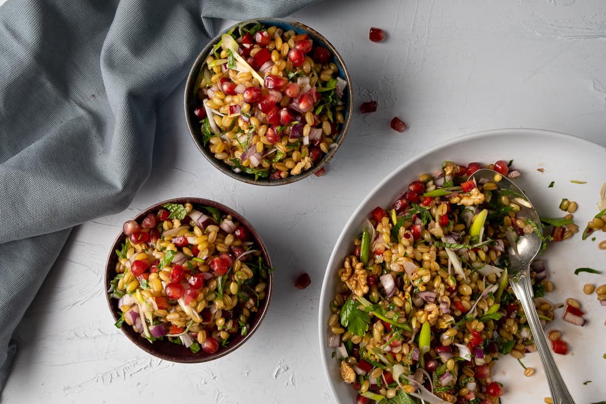 Salad with wheat berry grain and herbs in a white serving bowl and served in two black bowls and a grey napkin on the side.