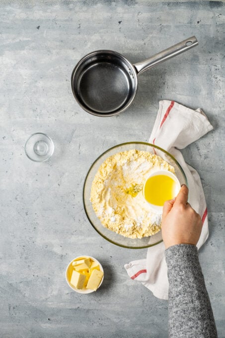 A woman's hand is pouring oil into a large bowl with cornmeal on a grey background.