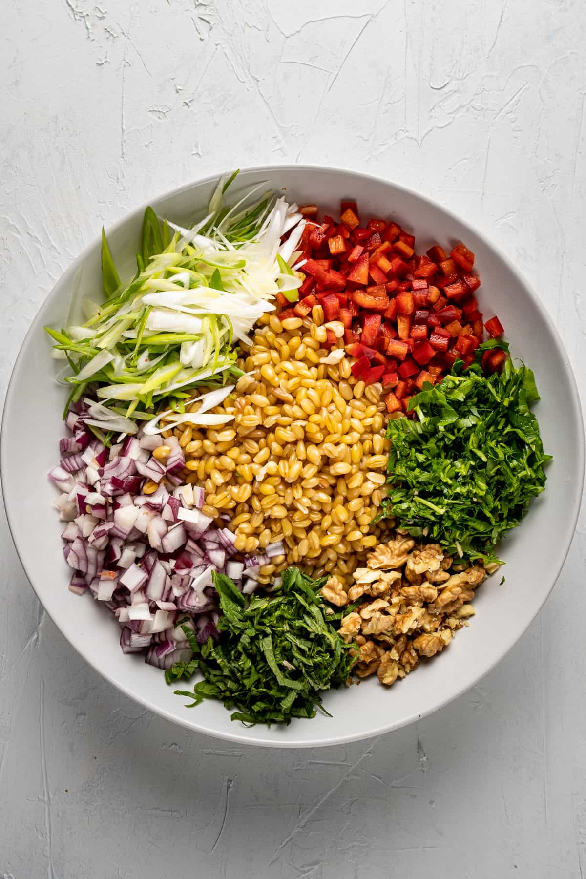 Cooked wheat berries, diced red onions and sweet red peppers, chopped herbs and walnuts arranged in a white bowl.