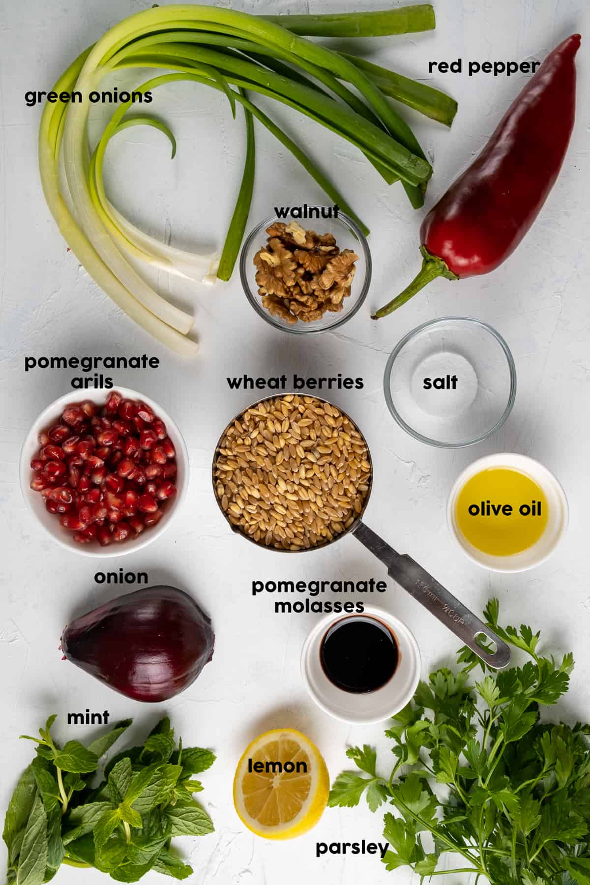 Green onions, one sweet red pepper, fresh parsley and mint, wheat berries, red onion, olive oil, pomegranate molasses, half lemon, pomegranate arils on a light background.