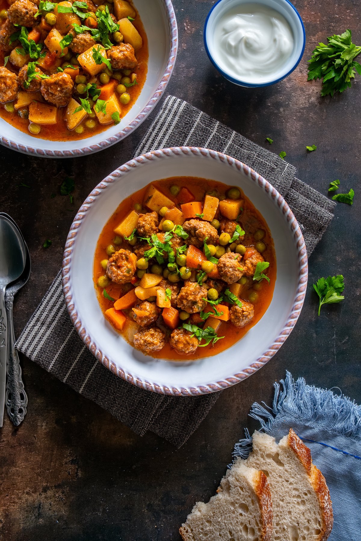 Two bowls of Turkish Meatball Stew (Sebzeli Köfte) with peas, potatoes, and carrots in tomato sauce, served with yogurt and slices of bread on the side.
