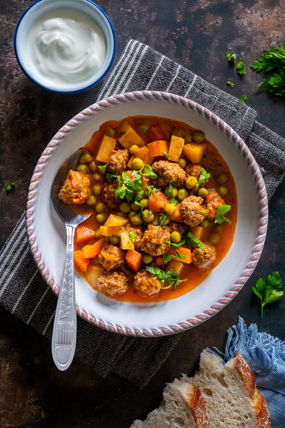 A bowl of Turkish Meatball Stew (Sebzeli Köfte) with peas, carrots, and potatoes in tomato sauce, served with yogurt and slices of bread.