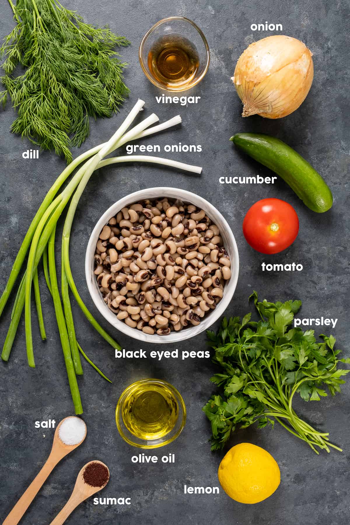 Black eyed peas in a white bowl in the middle and green onions, fresh dill and parsley, lemon, onion, cucumber, tomato, vinegar, salt and sumac around it on a dark background.