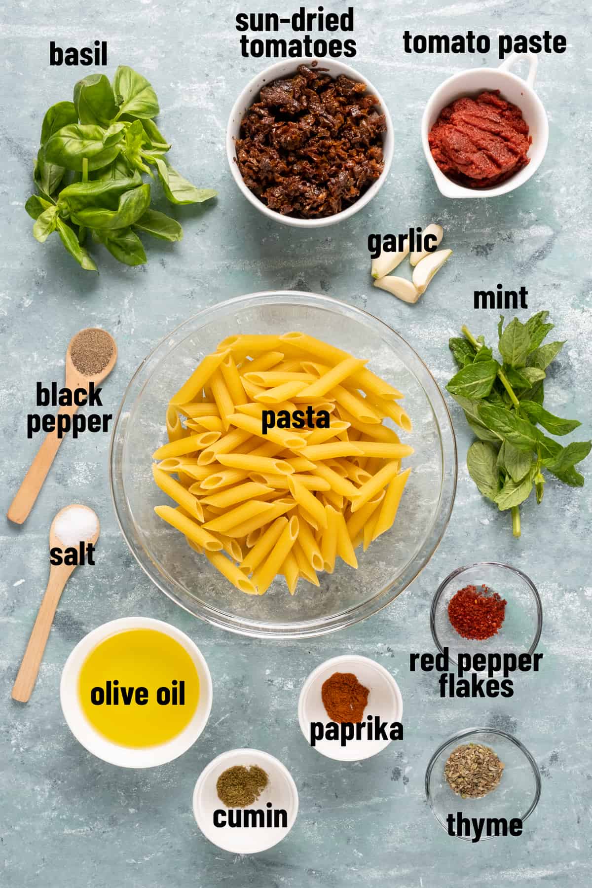 Penne pasta in a glass bowl in the center, sun-dried tomatoes, tomato paste, olive oil, spices, basil and mint leaves are around it on a grey background.