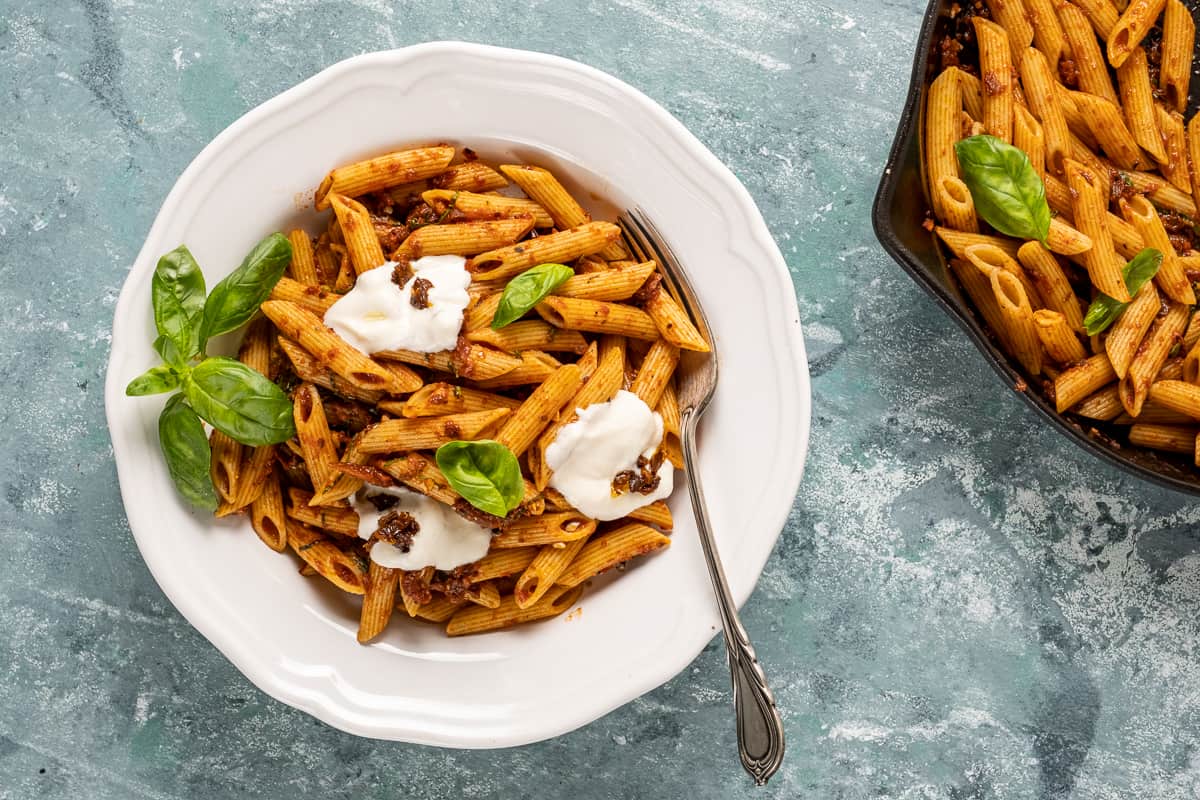 Sun-dried tomato sauce pasta in a white bowl topped with yogurt and basil leaves and more pasta in a cast iron skillet on the side.