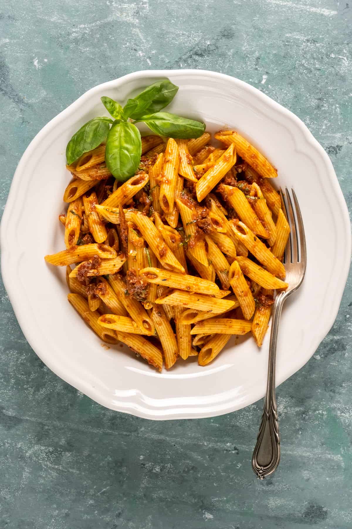 Pasta with a sun-dried tomato sauce in a white bowl topped with basil leaves and a fork inside.