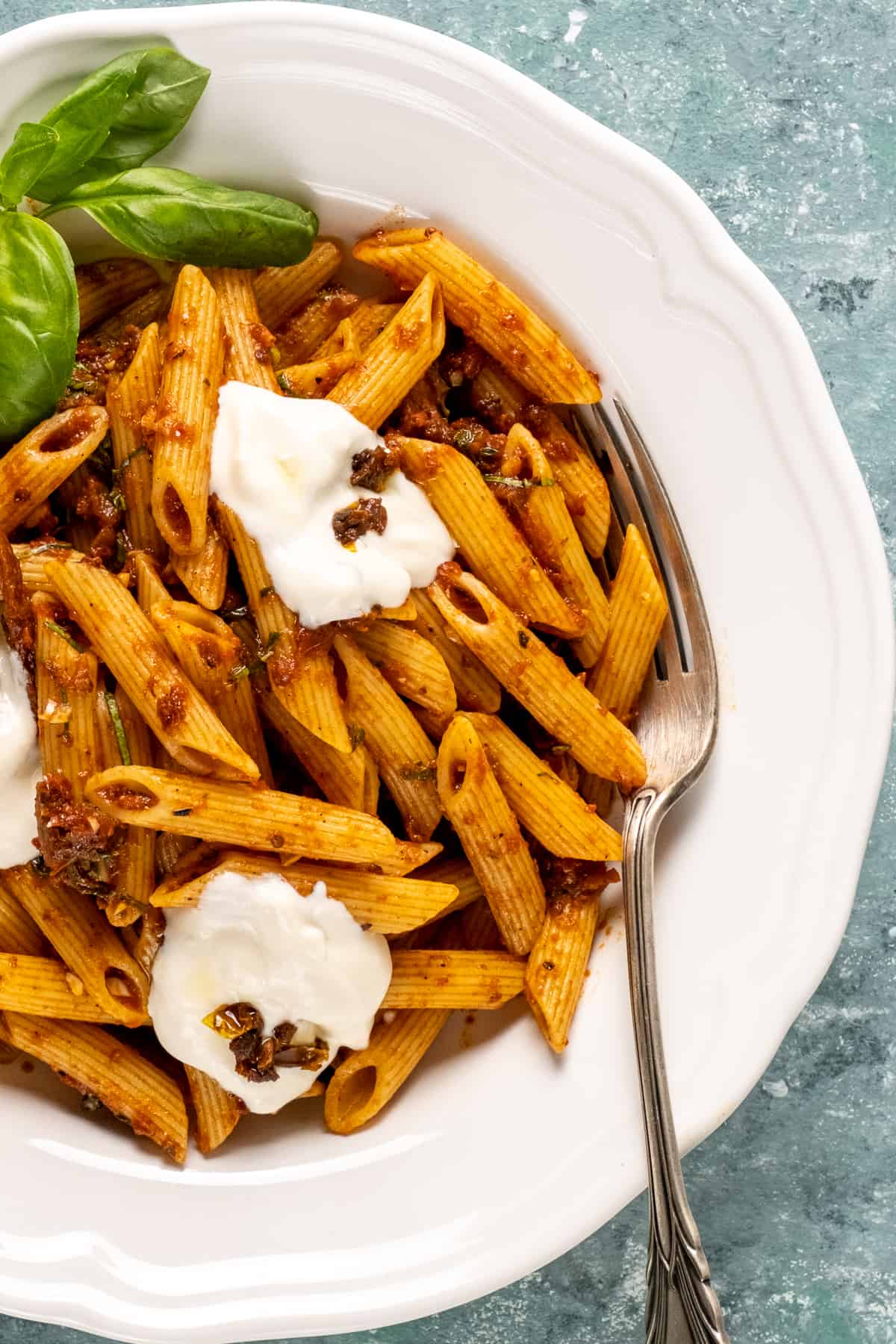 Sun-dried tomato pasta with basil and yogurt topping in a white bowl with a fork in it.