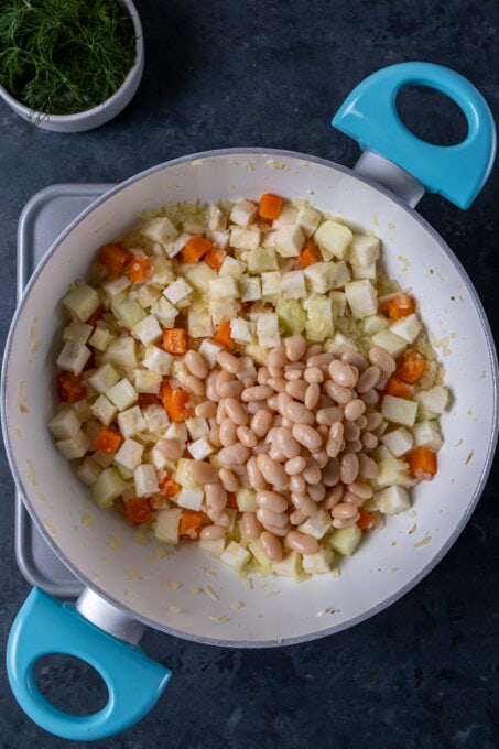 Cooked beans added over diced celery roots, carrots and apples in a white pan.