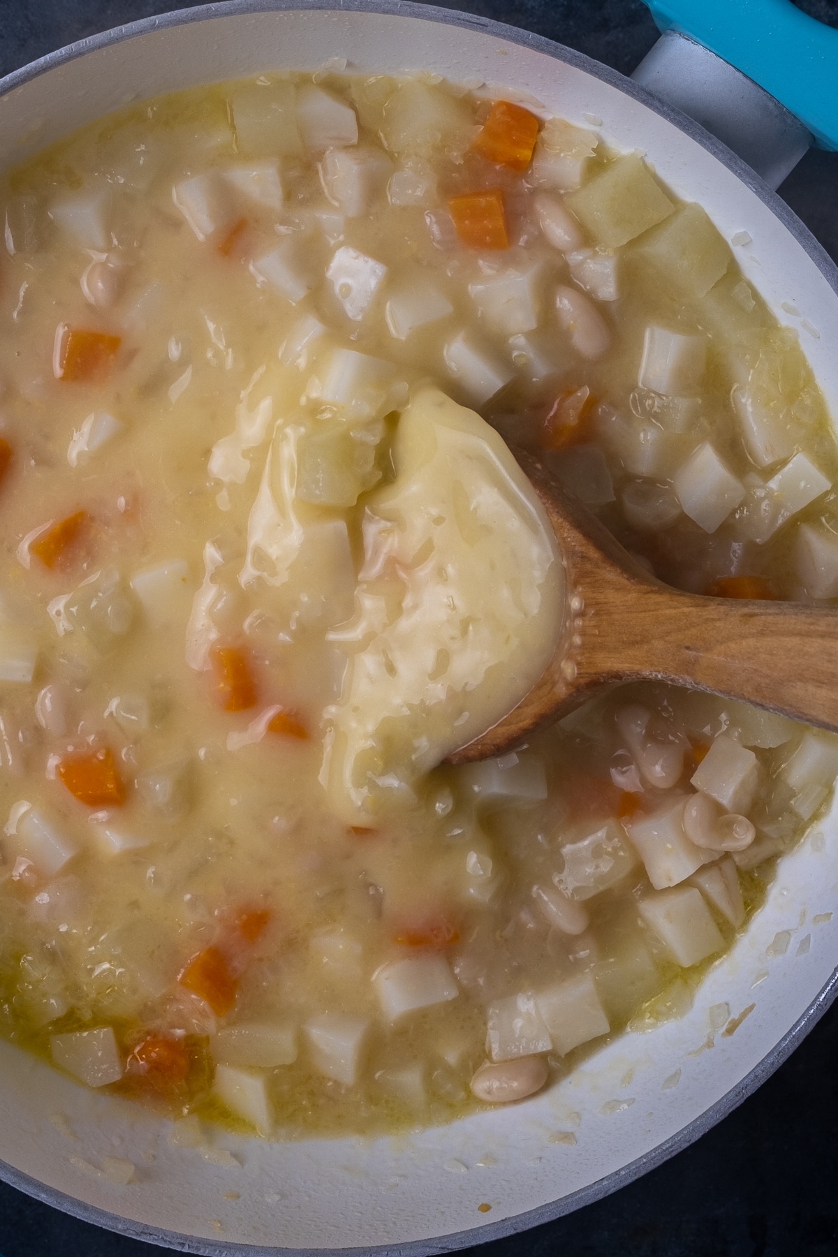 Celeriac and apple soup with beans in a white pan and a wooden spoon inside it.