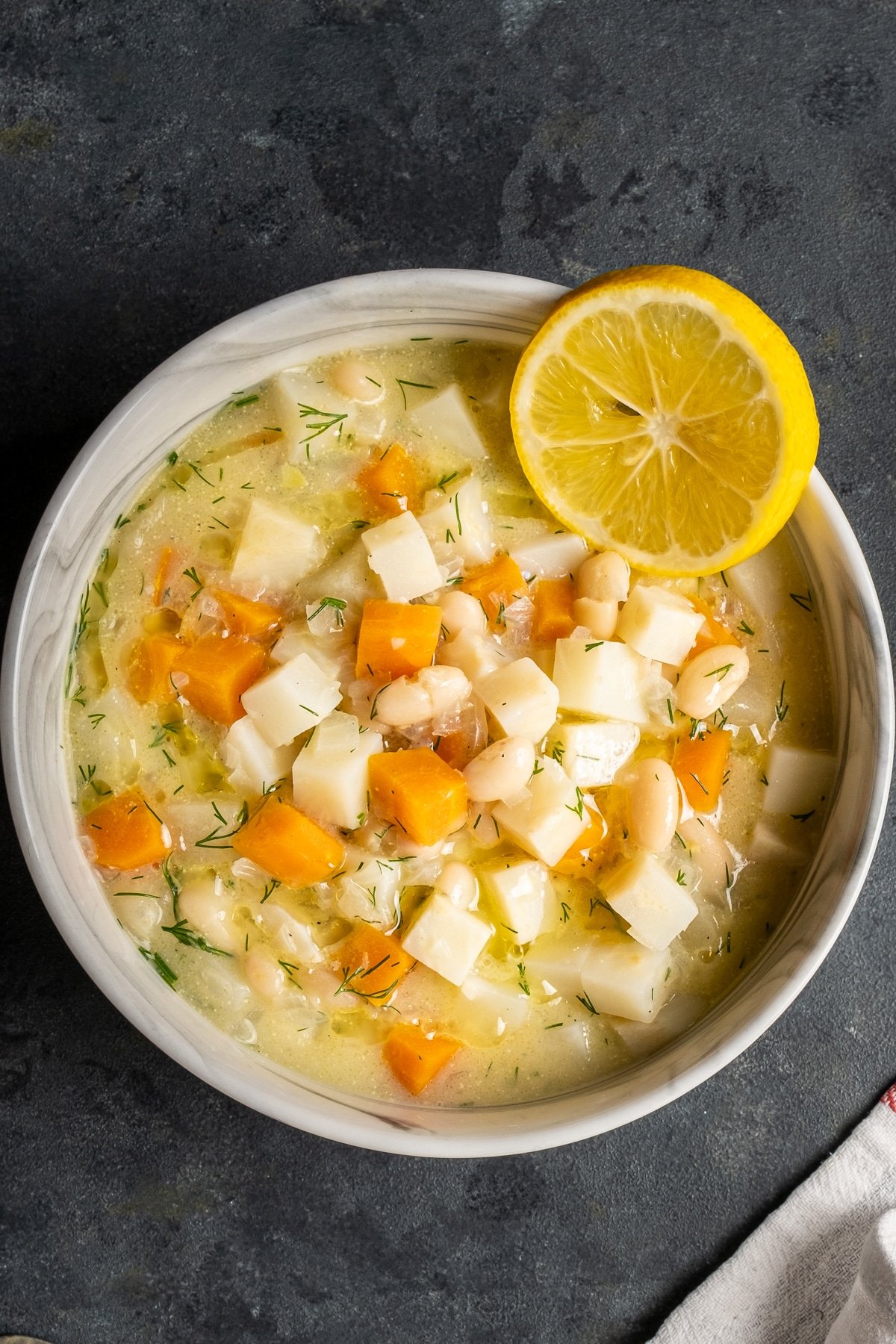 Soup with celeriac, apples, carrots and beans in a white bowl and a lemon slice on the side of the bowl.