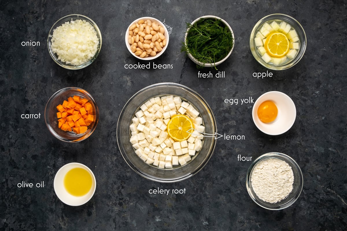 Diced celery root in a large glass bowl with water and lemon slice, diced apples, carrots, onions in separate bowls and cooked beans, egg yolk, flour, chopped fresh dill, olive oil in small bowls on a dark background.