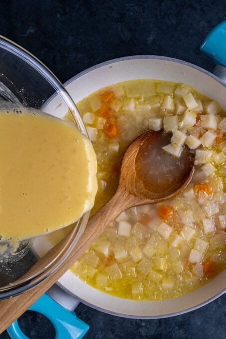 Pouring egg yolk lemon sauce from a glass bowl into the simmering soup with celery root, apples, carrots and beans.