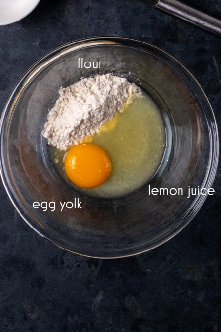 Egg yolk, flour and lemon juice in a glass bowl on a dark background.