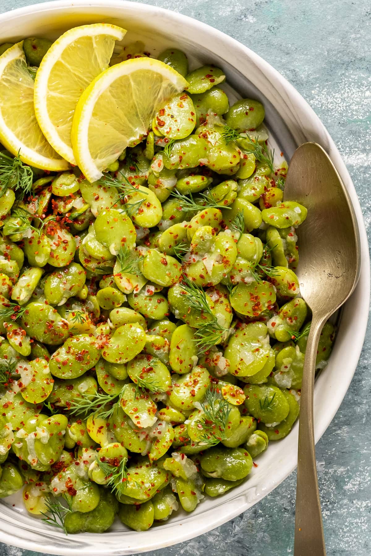 Fava beans cooked with onion and olive oil garnished with red pepper flakes, fresh dill and lemon wedges in a white bowl and a spoon in it.