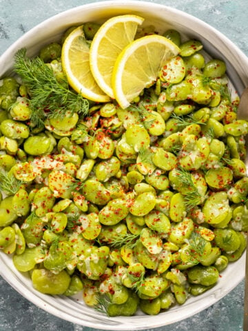 Fava beans cooked in olive oil garnished with red pepper flakes, fresh dill and lemon wedges in a white bowl and a spoon in it.