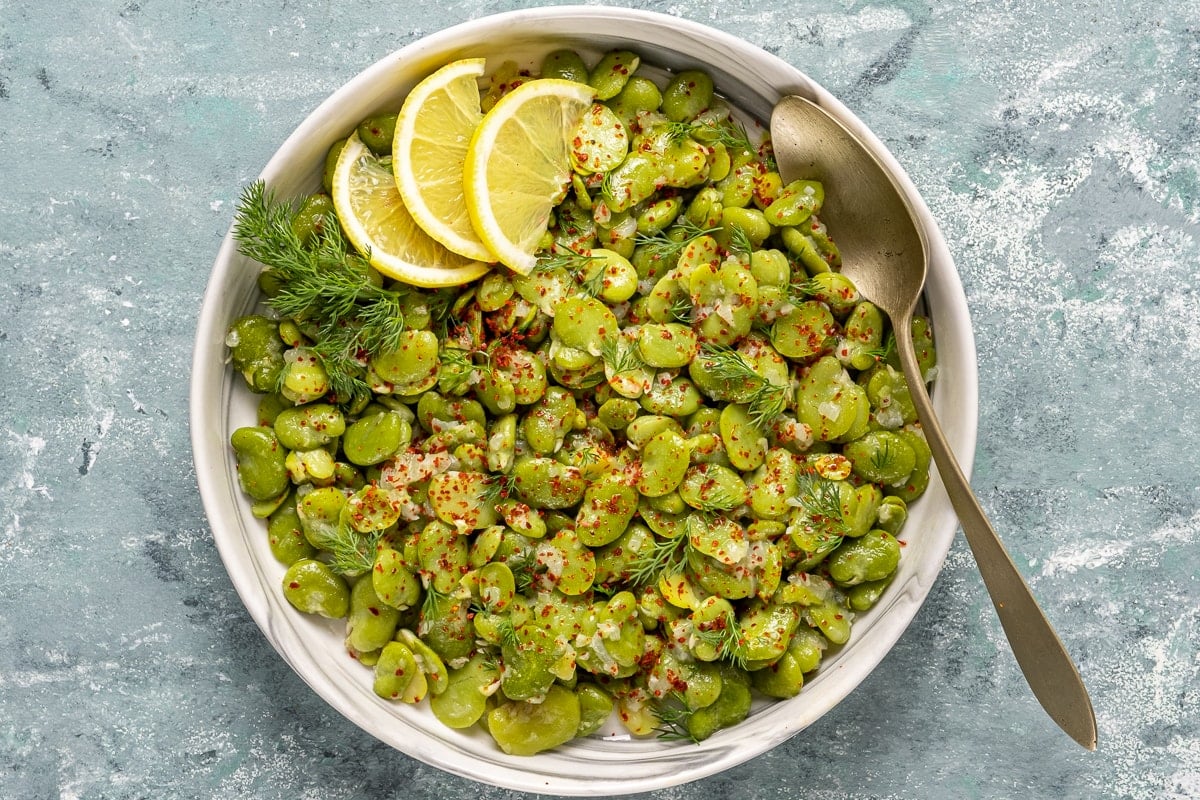 Fava beans topped with fresh dill, red pepper flakes and lemon wedges in a white bowl and a spoon in it.