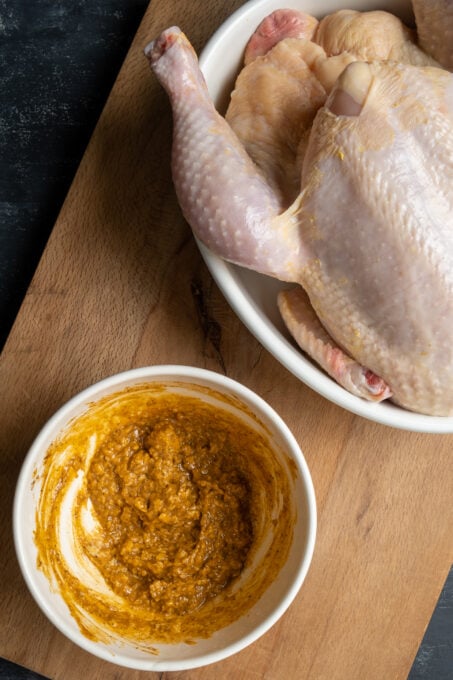 Marinade with butter and spices in a white bowl and a raw whole chicken in a pan on a wooden board.
