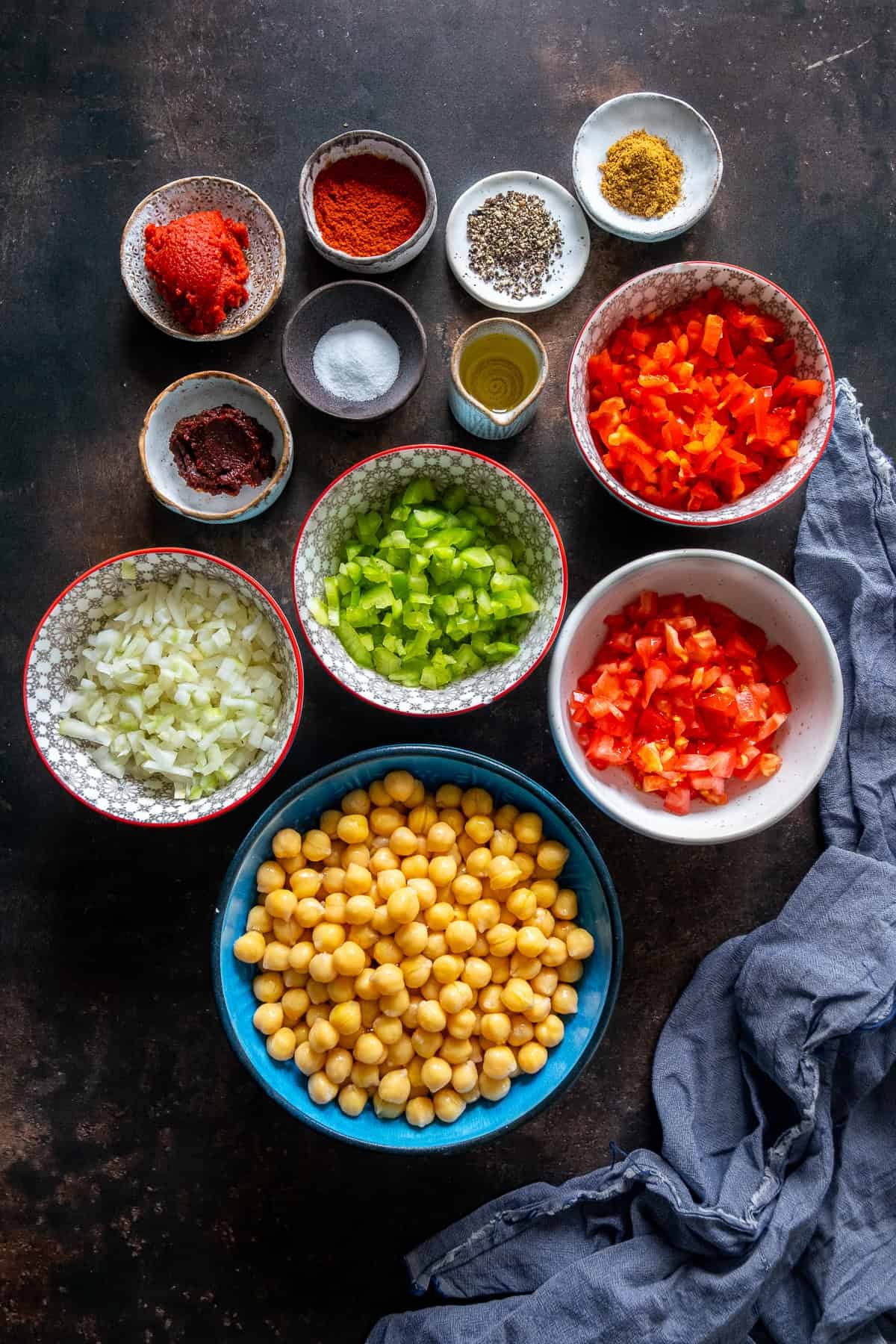 Ingredients for Turkish chickpea stew (Etsiz Nohut Yemeği) including chickpeas, diced onions, peppers, tomatoes, tomato paste, red pepper paste, spices, salt, and olive oil arranged in bowls on a dark background.