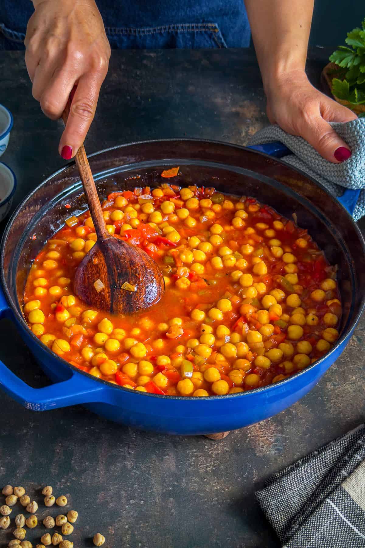 Turkish chickpea stew (Etsiz Nohut Yemeği) being stirred with a wooden spoon in a blue pot while simmering.