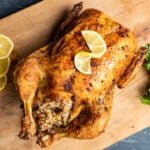 Rice stuffed whole chicken roasted in oven with a crispy golden skin photographed on a wooden board, lemon slices and a bunch of parsley on the side.