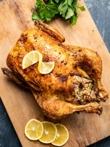 Roasted rice stuffed whole chicken on a wooden cutting board, lemon slices and a bunch of parsley on the side.