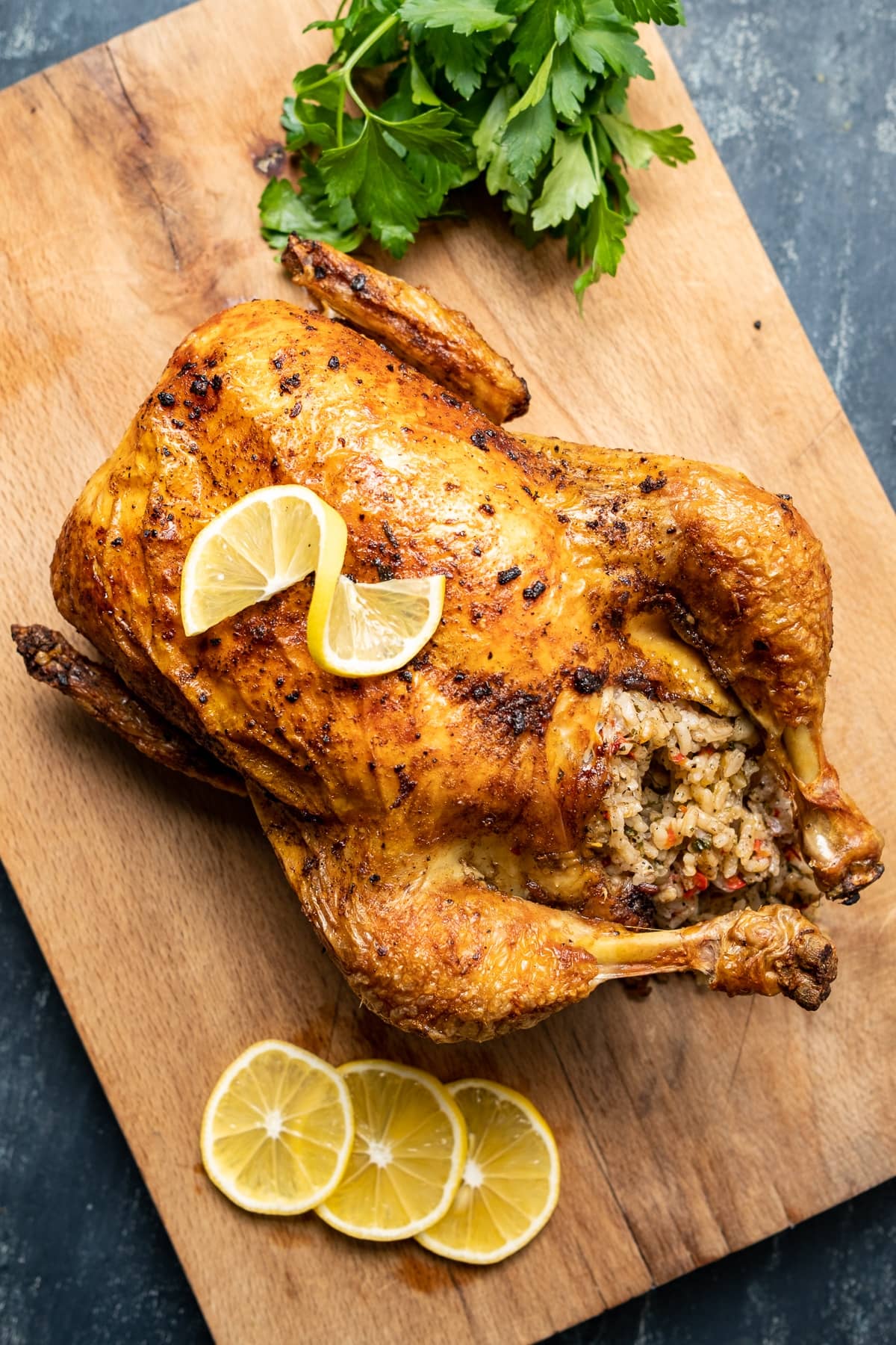 Roasted rice stuffed whole chicken on a wooden cutting board, lemon slices and a bunch of parsley on the side.