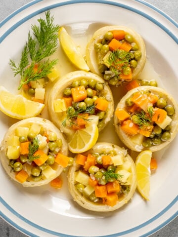 Artichoke bottoms stuffed with veggies like potatoes, peas and carrots garnished with fresh dill and lemon wedges on a white plate with blue stripes.