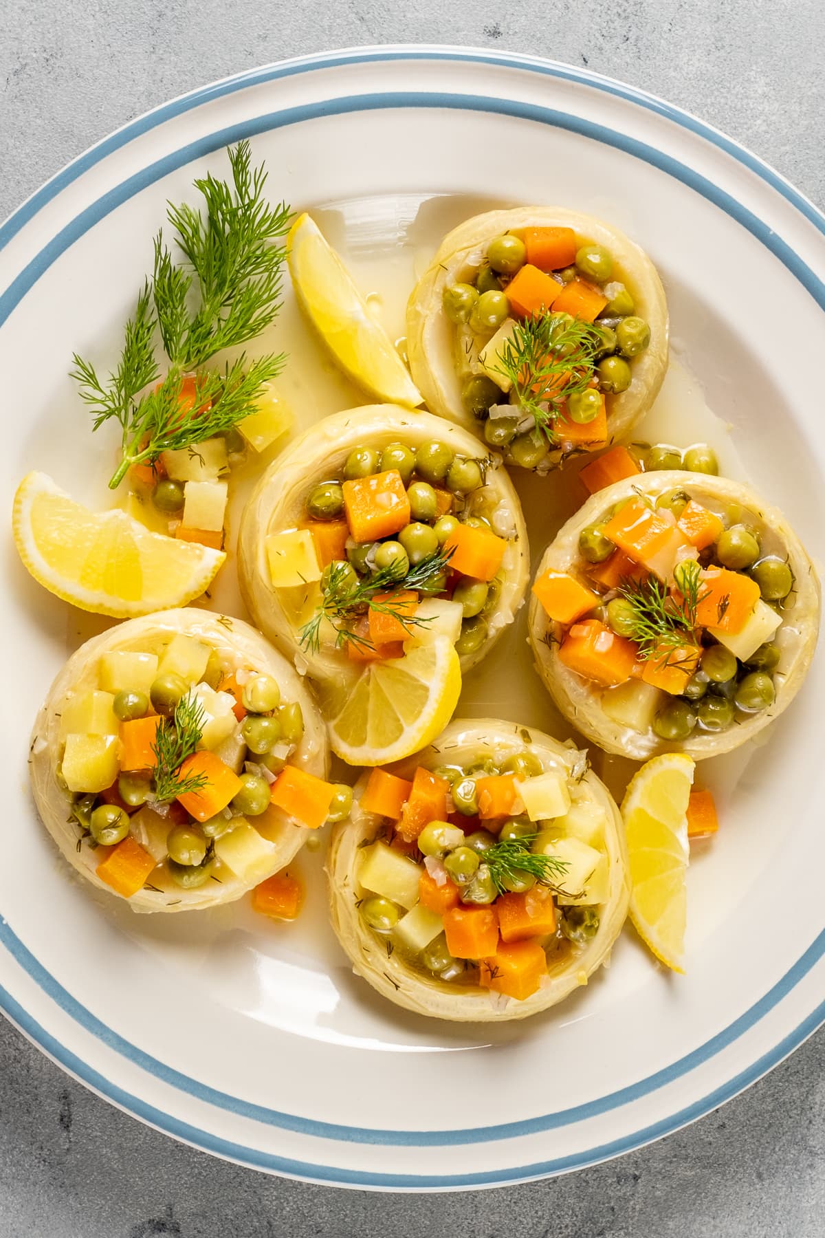 Artichoked bottoms stuffed with cooked peas, potatoes and carrots garnished with lemon wedges and fresh dill on a white plate with blue stripes.