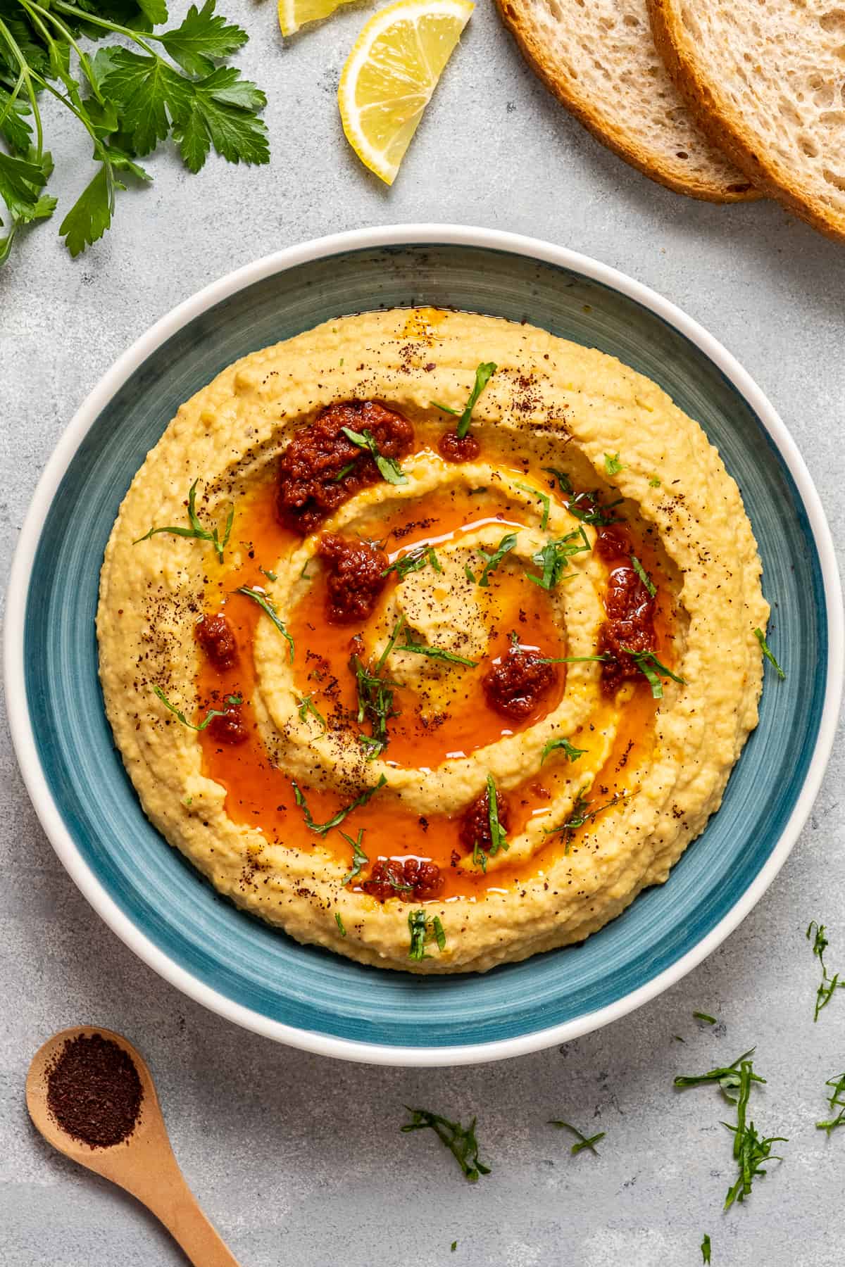 Lentil hummus topped with tomato paste oil sauce and minced parsley in a blue bowl and parsley, lemon slices, some bread and a teaspoon of sumac on the ground.