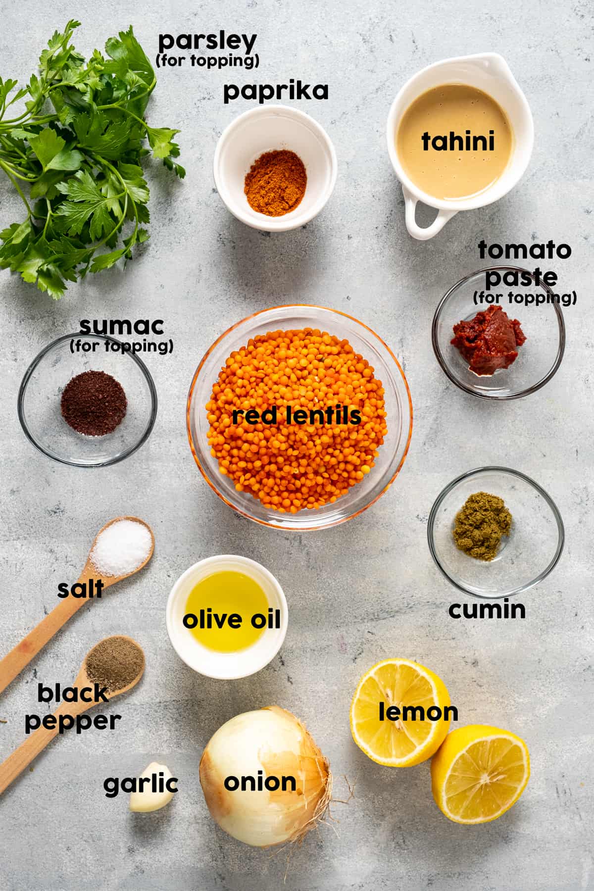 Red lentils, tahini, paprika, sumac, tomato paste, cumin, olive oil in small bowls and halved lemon, onion, garlic, salt and pepper and parsley on the ground.