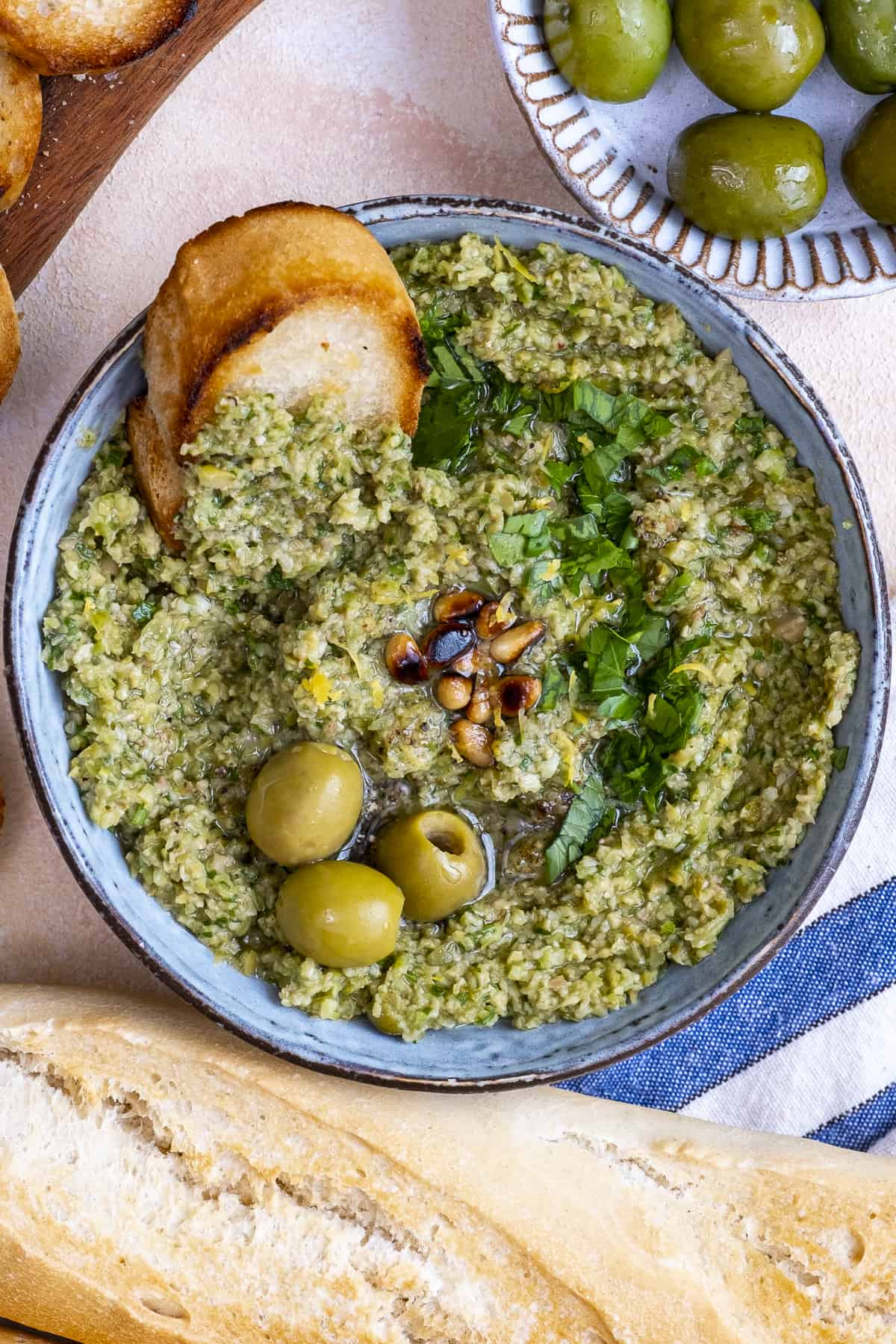 Green tapenade garnished with pine nuts, parsley and olives and a crostini dipped into it.