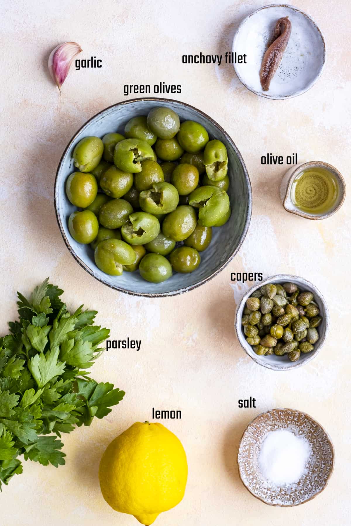 Green olives, capers, garlic clove, anchovy fillet, parsley, lemon, salt and olive oil in separate bowls.