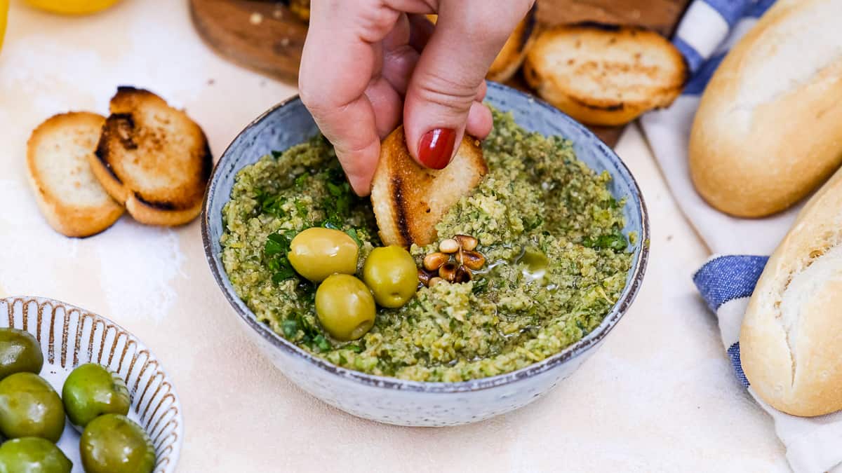 Hand dipping crostini into a bowl of green olive tapenade.