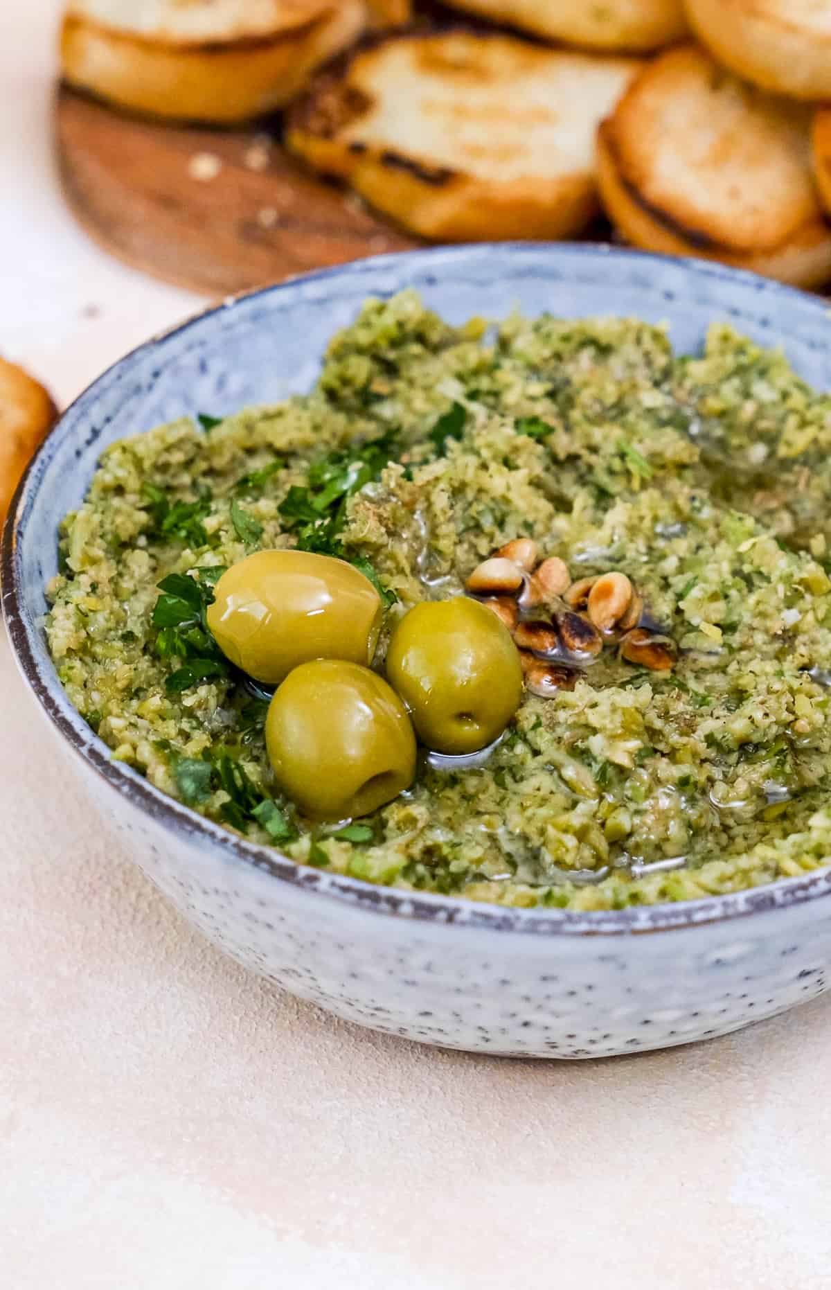 Green tapenade topped with olives and pine nuts in a bowl.