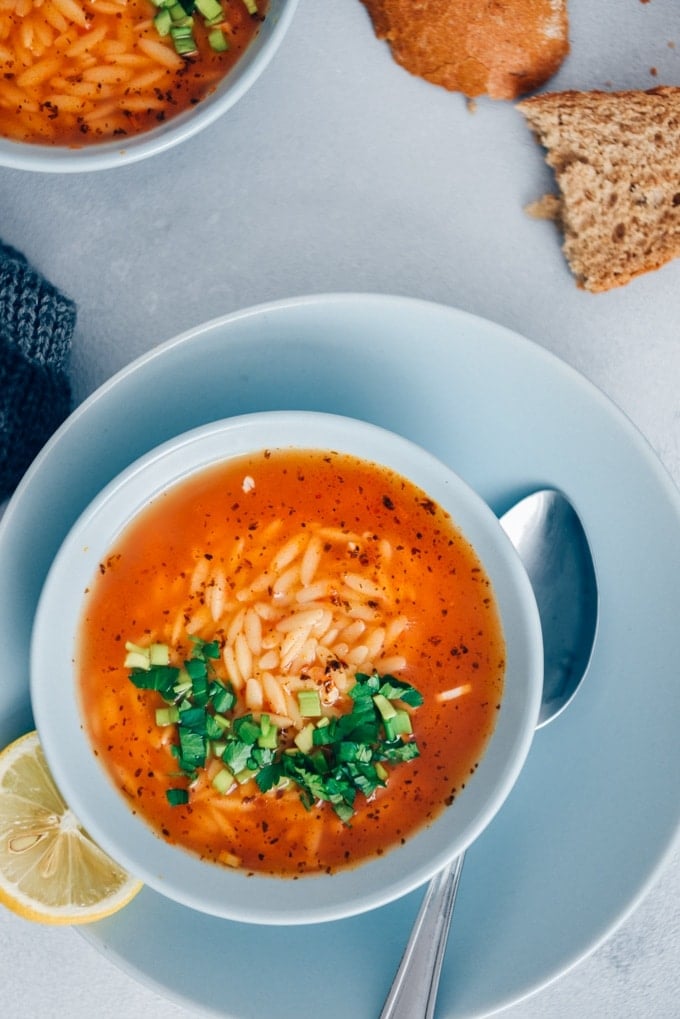 Turkish Tomato Orzo Soup Recipe packs a ton of flavor with fewer than 10 ingredients in just 20 minutes. The combination of tomatoes, lemon, parsley and orzo makes the easiest pasta soup ever. #orzosoup #wintersoup #souprecipe #turkishsoup Turkish tomato orzo soup garnished with parsley is served in light blue bowls on a grey background.